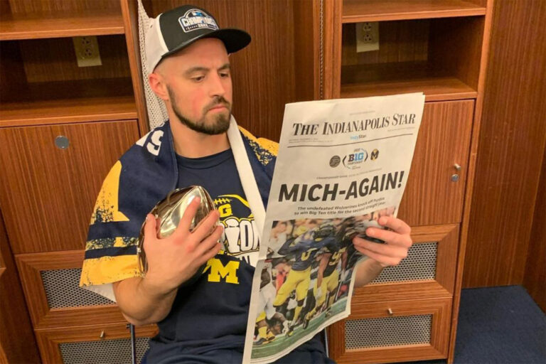 Connor Stalions in locker room posing with The Indianapolis Star newspaper and silver football trophy with headline reading "Mich-Again"